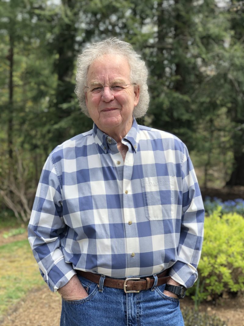 A photo of Brad Rose, an older white man wearing a blue and white plaid shirt and glasses. He has white hair and is looking into the camera.