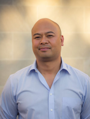 A headshot of Oliver de la Paz, a tan man wearing a blue shirt smiling and looking at the camera.