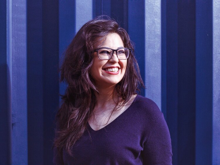 A Headshot of a smiling woman with long brown hair on a blue background looking to the left of the camera